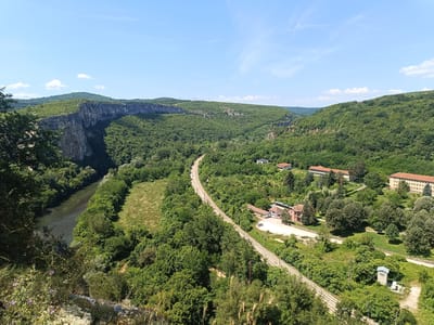 This is an aerial view of the Iskar Gorge in Karlukovo, Bulgaria. The image captures a winding railway line cutting through a lush, green valley dominated by a sheer, rugged cliff face on the left. A tranquil river flows alongside the cliff. Several buildings, appearing to be part of a village or settlement, are nestled amongst the trees on the right side of the valley. The sky is clear with a few scattered clouds, suggesting a bright, sunny day. The scene is a natural landscape with human infrastructure integrated into it.
