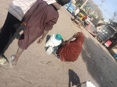 The image depicts an unpaved street scene in Gobirawa, Nigeria, under bright daylight. In the immediate foreground, two individuals are standing on dry, dusty ground, casting prominent shadows. One person on the left is partially visible, wearing a light-colored striped shirt, dark pants, and light-green sandals. Adjacent to them, another individual stands, seen from the back, wearing a long-sleeved brown garment and light-colored flip-flops.

To the right and slightly forward of the standing figures, a third individual is seated directly on the ground. This person is draped in a reddish-brown garment or hijab, with their right hand raised, covering their face. Next to the seated individual are a white bucket with a blue and green lid and a small, translucent white plastic container.

The background shows an unpaved road extending into the distance, bordered by various objects and structures. A yellow motorized tricycle (Keke Napep) is visible on the road, alongside several blue-colored vehicles. Scattered wooden structures, possibly market stalls or discarded items, are present. A reddish-patterned rug or mattress lies on the ground to the right. Utility poles with overhead wires and sparse trees are visible against a clear sky.