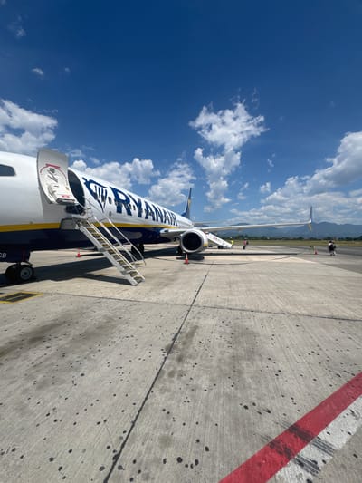 A Ryanair airplane is parked on the tarmac at the Tirana International Airport Nënë Tereza in Kruja, Albania. The plane's forward cargo door is open, and a set of mobile stairs is attached to the fuselage. The plane is white with blue and yellow trim and features the Ryanair logo on its side. The airport tarmac is concrete with tire marks and dark spots. In the background, a mountainous landscape is visible under a bright blue sky with scattered white clouds. Two individuals, a woman and a man, are visible in the distance near the plane, and another person is walking away from the plane.