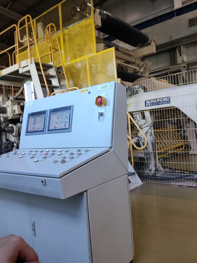 The image shows a control panel with two screens and numerous buttons in what appears to be an industrial setting in Shklov, Belarus. The control panel is light gray and features a red emergency stop button. The screens display data in tables and charts, suggesting they are monitoring an industrial process. Behind the control panel, there is yellow safety fencing and machinery, including large rollers. The overall environment is functional and industrial, with concrete floors and overhead lighting. The presence of industrial machinery and control systems indicates a manufacturing or processing facility.