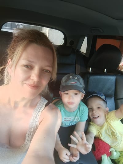 In a selfie taken from within a car, a woman with blonde hair pulled back and a light-colored lace-trimmed top smiles at the camera. To her right, two young boys are seated. The boy on the left, wearing a teal t-shirt and a grey shark-themed baseball cap, looks at the camera with a neutral expression. The boy on the right, in a yellow t-shirt and a blue baseball cap, grins widely, holding up his hand towards the camera. Behind them, the dark leather seats of the car are visible, with the interior of a building and a window with blinds visible through the car's rear window. The image appears to be taken during daylight, with sunlight casting a warm glow on the subjects. The specific location in Bender, Moldova, is not discernible from the image.