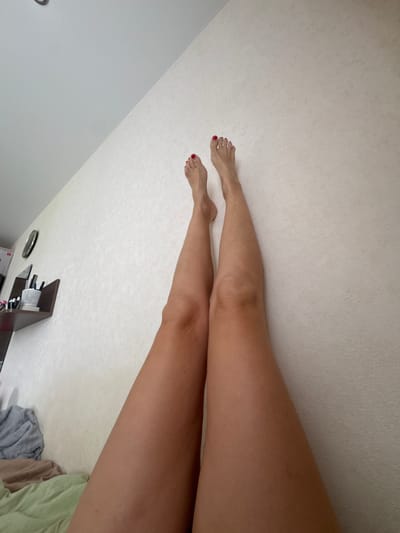 The image shows a view of a person's legs from a slightly downward-looking perspective. The person is lying on a bed with white patterned bedding, and the legs are stretched out towards the top left of the frame. The feet are visible, with red nail polish on the toes. A small shelf with various items is visible in the lower part of the image.
The location of the image is unknown. There is no information to indicate that the location of the image is in the city of Vesyoloye, Russia.
