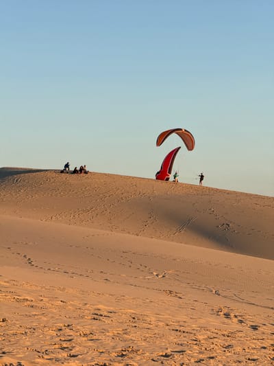 In the sand dunes of Lège-Cap-Ferret, France, at what appears to be dusk, several people are engaged in the activity of paragliding. Two large, colorful paragliders are visible, one partially inflated and leaning against a dune, the other in the air. One paraglider, predominantly red, has a person harnessed to it, preparing for flight. Another paraglider, orange and black, is higher in the sky. In the background, a group of individuals are seated or standing on a sand dune. The landscape is characterized by rolling sand dunes with subtle footprints and ripples, indicative of wind activity. The sky is a clear, soft blue, transitioning to a lighter hue near the horizon.