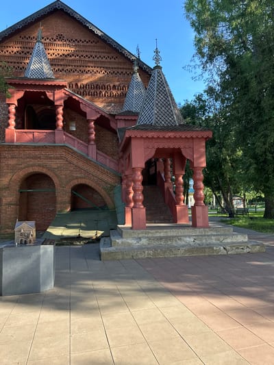 The image shows a traditional Russian wooden building with intricate red and grey ornamentation, set against a bright blue sky and lush green trees. The building features a steep roof with decorative gables and a prominent red staircase leading to an entrance. In the foreground, a tiled pathway casts shadows from the surrounding foliage. To the left, a dark grey pedestal displays a miniature architectural model, and a smaller, detailed model of a building sits on a stone base nearby. The scene appears to be a public space, possibly a historical site or museum, in the village of Terpilovo, Russia, given the architectural style and the presence of informational displays like the miniature models. The overall atmosphere is tranquil and sunny, suggesting it is daytime and likely during warmer months.