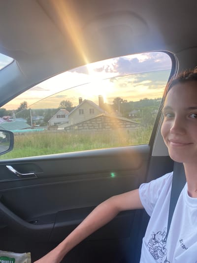 A young woman is taking a selfie inside a car, looking towards the right side of the frame with a smile. She is wearing a white t-shirt with a black graphic design on the left side. The car's interior is visible, including the door panel with a silver handle. Outside the car window, a rural landscape is visible during sunset. The sun's rays are strong, casting a golden glow and creating lens flare. Several houses with chimneys are scattered across a grassy hill. The sky is partly cloudy. The scene appears to be in the vicinity of Kurumoch, Russia, judging by the distinctive architecture and landscape typical of the region.