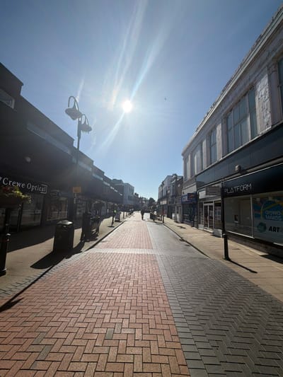 The image displays a pedestrianized street, Victoria Street, in Crewe, United Kingdom, on a bright, clear day. The sun, positioned high in the upper frame, generates significant lens flare and casts long, distinct shadows across the left side of the street. The sky is bright blue, featuring faint white contrails and a single bird in flight near the upper right corner.
The central part of the street is paved with interlocking red and grey bricks arranged in a herringbone pattern, bordered by grey slab sidewalks on both sides. On the left side of the street, a building with a dark facade houses "Crewe Optical," identified by its signage. Hanging flower baskets are suspended along this building. A black waste bin and a dark bench are situated on the left sidewalk. One individual, dressed in dark clothing, stands near the bin and bench, partially obscured by shadow. Ornate black street lamps with dual upward-curving light fixtures are present along this sidewalk.
On the right side of the street, an older, light-toned building features multiple windows on its upper story, showing signs of wear. The ground floor contains various shopfronts, including one marked "PLATFORM" and further down, "Coral." Two additional individuals are visible further down the street, walking away from the camera: one on the central paved pathway and another on the right sidewalk. No direct interactions between individuals are discernible.