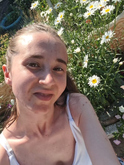 This is a selfie of a young woman lying down in a garden, surrounded by white daisies. She is wearing a white tank top. Her head is tilted, and she is looking towards the camera with a slight smile. The sun is shining brightly, casting shadows on her face and body. The background is filled with greenery and flowers, suggesting a warm, sunny day. There are also some hints of objects in the background, such as a blue object that could be a pot, and some gardening supplies. The overall mood is relaxed and peaceful. The woman's skin appears slightly tanned, and she has some freckles. The image is taken outdoors, likely in a backyard or park. The time of day appears to be daytime, judging by the bright sunlight and shadows. The location is Vypasne, Ukraine. There is no visible text in the image.