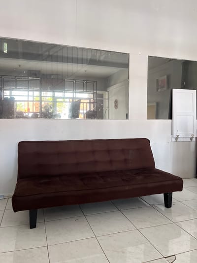 The image shows a dark brown futon sofa with tufted cushions, positioned centrally in the foreground on a tiled floor. The sofa has black legs and appears to be in a room with white walls. Above the sofa, a horizontal glass partition divides the space, revealing a reflection of a room with metal shelving units and some greenery outside. To the right of the futon, a white cabinet is mounted on the wall, with its doors partially open, revealing hinges. The overall environment appears to be an indoor space with a somewhat utilitarian aesthetic. There are no people or animals visible in the image. The location is identified as Kedawung, Indonesia.