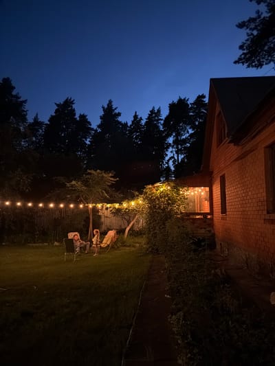 In the city of Udelnaya, Russia, at dusk, a brick house with a dark roof stands on the right side of the frame. The house's facade is illuminated by warm light emanating from its windows, which are visible through a covered porch area adorned with string lights. To the left of the house, a backyard is visible, featuring a patch of green grass. A wooden fence stretches across the backyard, also decorated with a string of lights. Two people are seated in lounge chairs on the grass, facing each other. One person, dressed in casual attire, is reclined in a chair, while the other, a child, stands beside a separate lounge chair. The background is dominated by a dense forest of pine trees, silhouetted against the deep blue twilight sky.