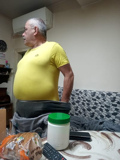 The image depicts an indoor scene, likely a living room or bedroom, with a man wearing a yellow t-shirt and grey undergarments in the process of dressing or undressing. He is standing and his midsection is prominently visible. In the foreground, on a patterned tablecloth, are several objects including a loaf of bread in plastic packaging, a white plastic container with a green lid, a mobile phone, and a remote control. A couch with a grey and white patterned fabric is visible in the background, along with an air conditioning unit mounted on the wall. The overall lighting suggests an indoor environment, possibly during the day or evening. The location is stated as Lomets, Bulgaria, though there are no visual cues in the image to confirm this specific location.