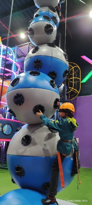 A child wearing a helmet and safety harness navigates a suspended obstacle course. The course consists of a series of large, blue and grey spheres connected in a line. The child is using handholds on the spheres to move across them. The background includes various other climbing structures and colorful lighting, suggesting an indoor adventure or amusement park setting.