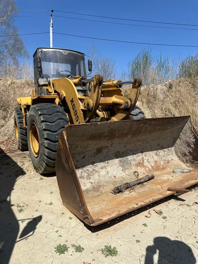 A front-loader heavy machine, possibly a tractor, is positioned on cracked, dry dirt. The machine is mostly yellow, with black tires. A large, rusty scoop or bucket is prominent. The side of the machine bears the inscription "XG951". A power line is in the background, with some dry grass and trees. The shadow of a person holding a camera is cast on the dirt. There is nothing in the image to confirm the location is Sadovoe, Russia.