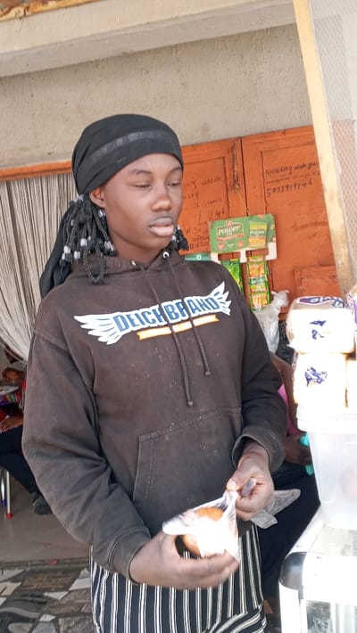 The image depicts an individual standing in what is identified as a commercial area in Dakata, Nigeria. The central figure is a young person with dark skin, wearing a black head wrap, a dark-colored hoodie with a white "DEICH BRAND" logo featuring wings on the chest, and black and white vertically striped lower garments. Braided hair with light-colored beads is visible on the left side of the head. The individual's eyes are closed or directed downward, and both hands are manipulating a clear plastic bag containing a small, round, light-brown item.
In the background, an orange-brown wooden panel, resembling a door or wall segment, is marked with handwritten text including "MONDAY" and the partial number "503399746". A green sign displaying "POWER OIL" is affixed to this panel, with several small, colorful packaged goods hanging beneath it. To the right of the central figure, a white plastic container holds multiple stacks of white rectangular packages with blue markings, consistent with packaged bread or similar retail items, along with a white plastic bag. To the far left, a white curtain partially obscures a seated individual and a metal chair leg. The floor exhibits a patterned tile design in black, grey, and white.