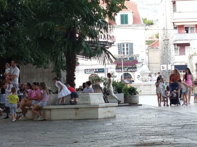 In Trebinje, Bosnia and Herzegovina, an outdoor scene depicts a plaza with several people, primarily children, engaged in various activities. A stone bench serves as a focal point, with a group of girls sitting on it. One girl in a white dress stands near the bench, looking towards a stroller. A man is holding a baby. The background features buildings with traditional architecture and greenery. The ground is paved with cobblestones. The overall atmosphere suggests a public gathering or leisurely outing.