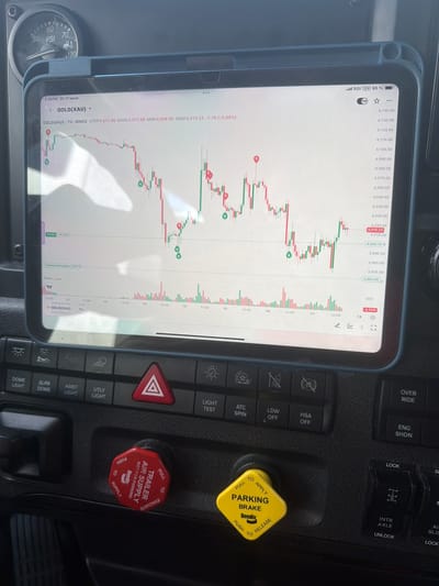 The image shows the interior of a truck cab, with a tablet computer mounted on the dashboard displaying a financial chart. The tablet displays a graph of GOLD(XAU) with timestamps and price indicators. Below the tablet, there are various truck control buttons, including a red emergency brake lever and a yellow parking brake lever. A PSI gauge is visible on the top left of the dashboard. The overall scene suggests the truck is stationary, possibly parked. The location is identified as Lebanon, United States of America.