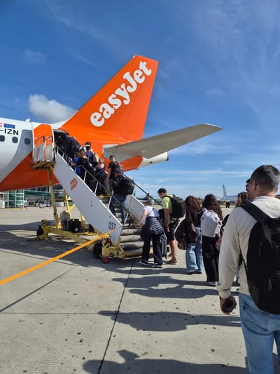 Passengers are boarding an EasyJet airplane at a tarmac in Moreira, Portugal. The plane has an orange tail with the EasyJet logo, and the mobile stairs are in place for boarding. People are walking up the stairs, and one person is standing on the tarmac, near the stairs. The sky is blue and sunny.