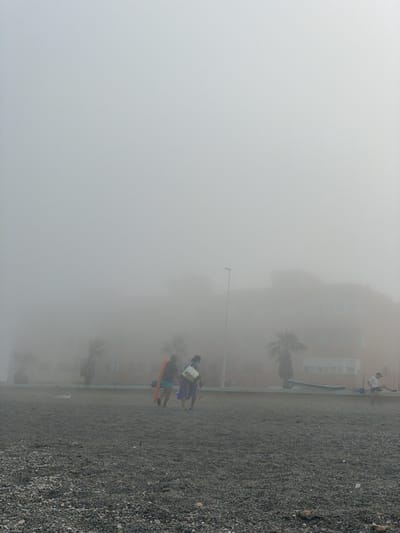 The image depicts a hazy beach scene with a gravely shore. The fog is thick, obscuring much of the background, but glimpses of buildings and palm trees can be seen through the mist. In the foreground, two people are visible, one carrying a brightly colored object, possibly a surfboard or bodyboard, and another person is partially obscured behind them. Further in the background, another individual is seen standing, facing away from the camera, with a building behind them. The weather appears to be foggy and possibly damp, which could suggest an early morning or overcast day. The presence of a beach, buildings, and palm trees indicates a coastal or resort-like setting. The activity taking place seems to be casual beach-goers in transit, perhaps heading to or from the water, or simply strolling along the shore. The fog creates a somewhat muted and serene atmosphere, with the limited visibility contributing to a sense of mystery or quietude. No text is visible in the image.