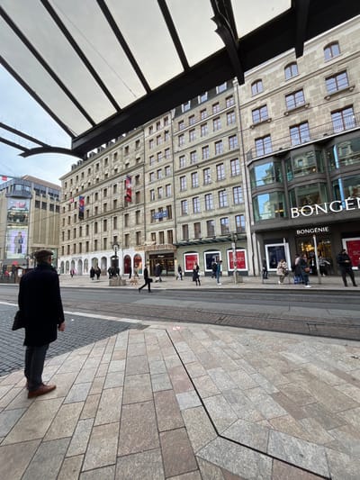 The image captures an urban street scene in Genève, Switzerland, rotated 90 degrees clockwise from its natural orientation. It depicts a wide paved area in the foreground and right, transitioning to a tram track and a row of large buildings on the left and in the background.
**Subjects:** Several people are visible throughout the frame, walking or standing along the street and near the storefronts. In the immediate foreground, a person in a dark coat and brown shoes walks away from the viewer. The prominent building on the left features a classic stone facade with numerous rectangular windows on upper floors and a modern glass and dark-panelled ground floor housing a department store. A transparent or frosted shelter-like structure, possibly a tram stop, partially occupies the far left foreground.
**Activity:** People appear to be engaged in everyday urban activities such as walking, window shopping, or waiting. The general atmosphere suggests a typical daytime city environment.
**Notable details:** The time of day appears to be daytime, under an overcast sky, as indicated by the diffused light and lack of strong shadows. The street features tram tracks, suggesting it's a route for public transportation. The paving stones vary in shade, creating a visual pattern on the ground. The architecture is characteristic of a European city center, with multi-story buildings.
**Visible text:** The most prominent text is "BONGÉNIE," displayed vertically on the modern section of the large building. Other partially visible text includes "SOLEILDES" on a display sign, likely "Soleil des..." for a brand or product.