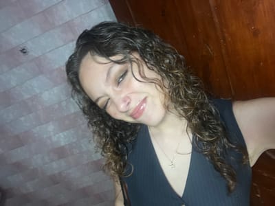A young woman with curly brown hair and fair skin is smiling towards the camera. She has a nose piercing and is wearing a navy pinstripe vest and a delicate necklace. The background consists of two different walls, one with a light-colored striped pattern and the other a darker brown wood paneling. The lighting is somewhat dim, giving the image a casual, indoor setting.