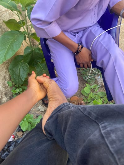 The image shows an outdoor scene in Nagazi, Nigeria. A person wearing a lavender-colored outfit sits on a blue plastic chair. The person has a beaded bracelet on their left wrist and is holding a white cable. Next to them, another person wearing black jeans is seated with their right foot visible. A hand with red nail polish touches the foot. Green plants and a concrete surface are visible in the background.