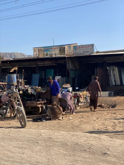This image depicts an outdoor commercial scene in کوکران, Afghanistan. The sky is clear blue, with a rugged mountain range visible in the distant background. The ground is unpaved, consisting of dusty, uneven dirt.
In the foreground, a reddish-brown motorcycle is parked on the left. Adjacent to it is a functional two-wheeled trailer loaded with various items, including a blue barrel and dark metallic objects, suggesting a workshop setup.
Four adult males are present. One male, wearing a blue jacket over light-colored trousers, stands near the trailer, looking downwards at the ground. To his right, another male, dressed in a dark tunic and trousers, is seated on a metal chair, facing away from the viewer. A third male, clad in a brown shawl, is crouched on the ground directly in front of the seated male. These three individuals appear to be interacting with the workspace around the trailer. A fourth male, also dressed in a dark brown tunic and trousers, is walking towards the right side of the frame, away from the central group. A second, covered motorcycle is parked behind the seated man.
In the midground, a row of single-story shops is visible, featuring corrugated metal roofs and open fronts. Two prominent signs are mounted above these shops. The left sign reads "شادی فلزکاری" (Shadi Metalwork), and the right sign reads "غلام رضا فلزکاری" (Gholam Reza Metalwork), indicating that the establishments specialize in metal fabrication or steelwork. Various metal sheets, frames, and other metal products are visible within and outside the shop entrances. The scene is brightly illuminated by direct sunlight, casting distinct shadows.