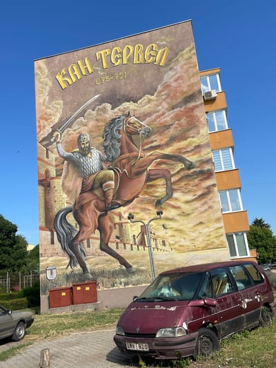 This is a photograph of a mural on the side of a building in the city of Tervel, Bulgaria. The mural depicts a historical figure, identified by the text above as "КАН ТЕРВЕЛ" (Khan Tervel), a Bulgarian ruler from 675-721. The figure is shown on horseback, raising a sword. The background of the mural features clouds and a castle. In the foreground, two red electrical boxes are visible. A dark red minivan is parked in front of the building, with its paint peeling and vegetation growing around it. To the left of the minivan, part of a light-colored car is visible. The scene is set on a paved area with grass and trees surrounding it. The sky is clear and blue.
