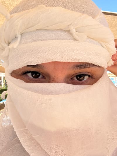 The image presents a close-up, upward-angled view of a person whose face is largely enveloped by a voluminous white, loosely woven fabric, resembling a scarf or head covering. Only the person's eyes, dark eyebrows, and a small portion of the upper face and nose are visible through an opening in the textile. The eyes are dark brown with prominent, long eyelashes, and they gaze directly at the camera. A hand, likely the subject's, is visible at the top of the frame, appearing to adjust or hold the fabric around the head.

The white fabric is a dominant feature, exhibiting a distinct texture with a grid-like, open weave, suggesting a lightweight material such as cotton or linen gauze. It is artfully draped, showing folds, knots, and frayed edges, creating a dynamic visual.

The background is softly blurred but reveals elements consistent with an outdoor daytime setting. A clear blue sky is visible at the top, along with hints of light-colored architectural structures or walls. In the lower right, glimpses of green foliage or trees can be seen. The bright, natural light illuminates the scene evenly.

The activity captured is a direct, engaging look from the hidden person. Given the provided context, the setting is Safaga, Egypt, and the attire aligns with traditional or protective headwear in warm, sunny climates. There is no visible text in the image.