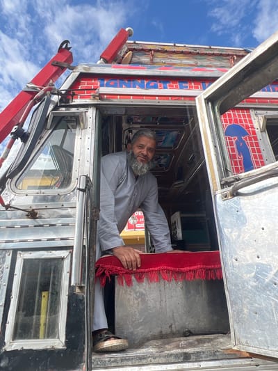 A man with a beard and gray shirt is standing in the open doorway of a truck in Bodhkharboo, India. The truck is decorated with red, blue, and white lettering and colorful designs. The man is leaning on a red fringed cloth that covers the top of a metal compartment. The truck has chrome accents and windows. The sky is blue with white clouds.
