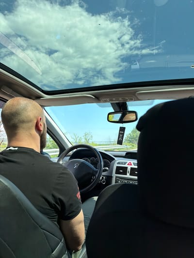 The image depicts the interior of a Peugeot car, likely in Belovitsa, Bulgaria. The view is from the passenger seat, angled towards the driver. The driver, a bald man with a black T-shirt, is either resting or asleep, as he is reclined towards the center console. The steering wheel prominently features the Peugeot logo. The car's dashboard and console are visible, along with an air freshener hanging from the rearview mirror. The sunroof is open, reflecting a cloudy sky. Outside, there are trees and greenery. The scene suggests the car is parked on a road.