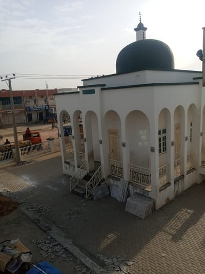 The image captures a mosque located in Hotoro North, Nigeria. The mosque features a prominent dark green dome topped with a crescent moon and star. The building is painted white with arched doorways and windows. A sign above an entrance reads "MOSQUE" in green lettering. In the foreground and surrounding areas, there is evidence of construction or renovation, with stacks of stone slabs, debris on the ground, and piles of dirt. Adjacent to the mosque, there are buildings with storefronts, including one labeled "RUFAIDAH YOGHURT & FURA" and another with "MDS MACY." Several people are visible in the scene, some near the storefronts and others engaged with a red truck that appears to be carrying goods. A utility pole with electrical wires stands to the left of the frame. The ground is paved with interlocking bricks in most areas, with some sections of rougher terrain where construction is occurring. The sky is clear and bright, suggesting daytime.
