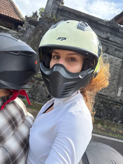 Two individuals are seated on a motorcycle in front of a Balinese temple structure. The person in the foreground, wearing a pale yellow helmet with a black face mask, is looking towards the camera. Their hair is curly and auburn. They are wearing a white jacket. The person behind them is wearing a black helmet and a plaid shirt. A red bandana is visible tied around their neck. The temple in the background has traditional Balinese architecture with a tiled roof and stone carvings. The sky is partly cloudy.