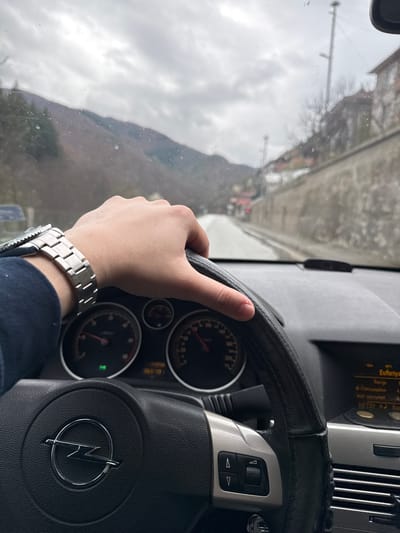 A person is driving a car, gripping the steering wheel with their left hand. The steering wheel features a logo, indicating the car is likely an Opel. The person is wearing a silver wristwatch on their left wrist. Outside the car window, a landscape with mountains and some buildings is visible. The location appears to be Byala Reka, Bulgaria. The weather seems cloudy or overcast. The road ahead appears to be a narrow street with buildings on one side and a mountainous landscape on the other.