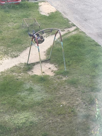 An overhead shot captures a person on a playground swing. The person, wearing dark clothing with a lighter colored shirt partially visible, is seated on a swing set. The swing set has a curved metal frame, painted in red, blue, and yellow, and a horizontal bar. To the left of the swing set is a metal climbing frame with horizontal bars. The ground consists of patchy green grass and sand. In the background, to the left, a paved surface with a curb is visible. The scene appears to be outdoors during the daytime, under natural light. There are no discernible emotions or specific weather conditions indicated, other than it appearing to be a clear day. No visible text is present in the image. The location context of Asipovichy, Belarus, suggests this is likely a public park or residential area playground.