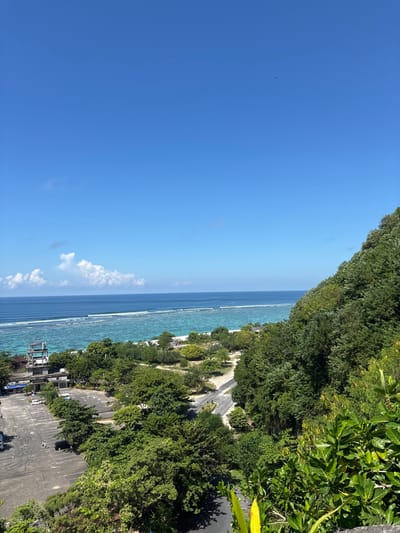 The image shows a coastline in Kutuh, Indonesia, from an elevated perspective. The scene features a deep blue sky with a few scattered clouds. A light blue ocean stretches alongside a coastal road with buildings and parking areas. Lush green trees cover the land bordering the road. A small bird is seen in the sky.