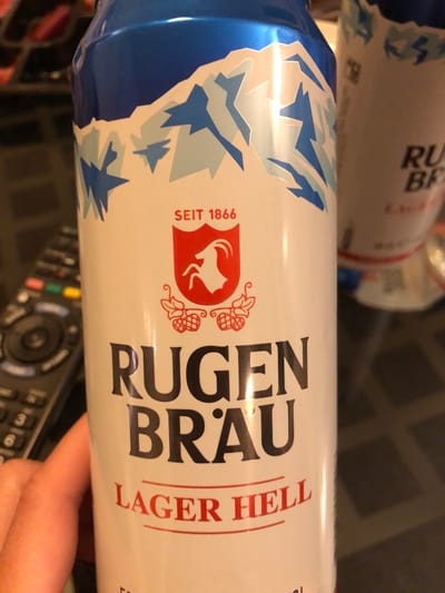 The image shows a white beer can with blue and white mountain designs near the top. The can features the brand name "RUGEN BRAU" in large black letters, with "LAGER HELL" in red below. A red emblem with a bull's head silhouette and the inscription "SEIT 1866" is visible. A remote control is present in the foreground, with a person's fingers partially visible. Another can is partially visible behind the main can. The scene is likely indoors. Based on the brand name, the location is probably somewhere close to Interlaken in the canton of Bern in Switzerland. Kehrsatz is some distance from Interlaken, and not likely to be related to this image.