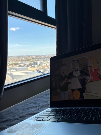 This media file depicts an indoor scene overlooking a city skyline. A laptop is open and positioned on a surface, likely a bed due to the quilted texture visible. The laptop screen displays an animated scene featuring characters from "Rick and Morty," specifically Summer, Rick, and Morty, engaged in conversation. A woman with blonde hair wearing a red dress stands, while Rick and Morty, in their signature green shirt and blue pants respectively, are seated.
The background of the laptop screen shows a bookshelf. To the right of the laptop, its keyboard is visible, with Thai characters on some of the keys indicating a Thai keyboard layout.
The laptop is placed near a large window, which offers a panoramic view of a densely populated city. The cityscape is characterized by numerous buildings, some appearing to be apartment complexes, and a train track cutting through the urban landscape. The sky is blue with a few scattered clouds, suggesting it is daytime. The angle of the sun, casting shadows and highlighting parts of the city, implies it is likely late morning or early afternoon.
The overall setting suggests a comfortable, perhaps residential, environment with a view of urban life. The presence of the laptop with a cartoon playing suggests a moment of leisure or relaxation. The time of day appears to be sunny and clear. The location is identified as Sao Thong Hin Town Municipality, Thailand, which aligns with the presence of Thai characters on the keyboard.