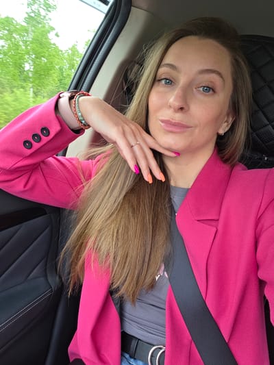 A woman with light brown hair and blue eyes is sitting in a car wearing a bright pink blazer, a gray top, and a seatbelt. She has bracelets on her left wrist and a ring on her left ring finger. Her nails are painted with alternating pink and orange polish. The background shows a green landscape visible through the car window. The interior of the car has black leather seats with quilted stitching. There is no indication that the location is Аур, Russia.