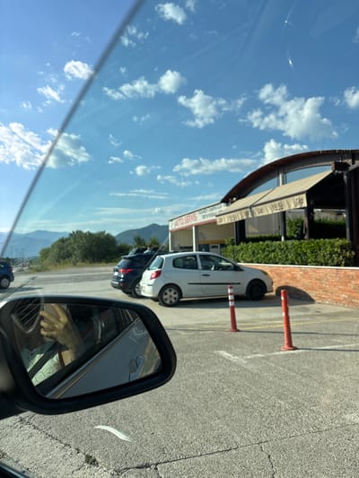 The image shows a view from inside a car, looking out towards a parking lot and a building. The sky is blue with white clouds. In the foreground, the car's side mirror reflects a person's hand holding a phone. In the parking lot, there are several cars, including a white hatchback and a dark SUV. There are also two orange traffic cones. The building in the background appears to be a cafe or restaurant with a modern design, featuring large windows and awnings. The text on the building signage is partially visible and appears to say "AUTO SERVIS". The overall scene suggests a sunny day in an urban or semi-urban environment. Based on visual cues such as architecture and landscape, the location is likely Dub, Montenegro.