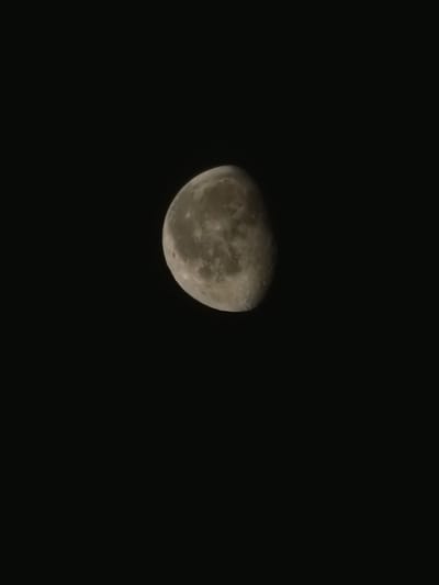 A close-up, vertically oriented photograph captures a waxing gibbous moon against a pitch-black night sky. The moon is positioned in the upper left quadrant of the frame, with its surface clearly illuminated and showing visible craters and maria. The rest of the image is devoid of any other celestial bodies or earthly objects. The scene suggests a clear night in Voluyak, Bulgaria, where the moon is a prominent feature in the absence of light pollution.