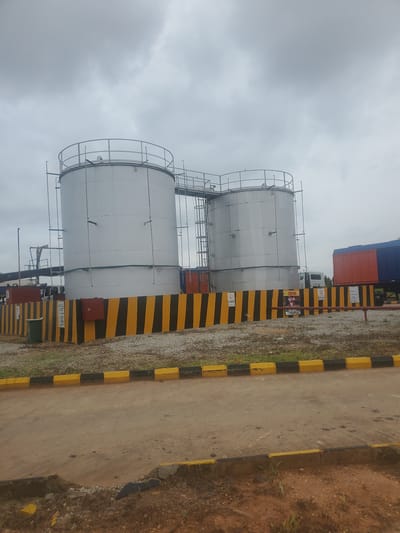 The image shows an industrial scene in Gwagwada, Nigeria. Two large, cylindrical storage tanks, painted silver, are prominently featured. These tanks are connected by a walkway and a ladder system. They are surrounded by a black and yellow striped barrier, indicating a hazardous or restricted area. A green garbage bin is visible near the left side of the tanks. Several caution signs are posted on the barrier. In the background, a semi-trailer truck with an orange and blue container is parked. The sky is overcast, suggesting a cloudy day. The ground around the tanks is covered with gravel and some sparse vegetation. A paved road or pathway is in the foreground, with yellow and black striped curbing.