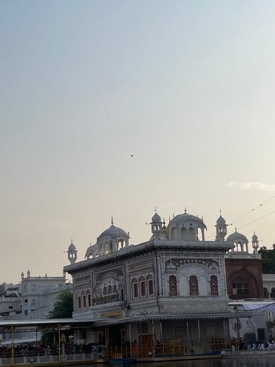 The image depicts the Harmandir Sahib, also known as the Golden Temple, in Amritsar, India. The structure is characterized by its white marble facade adorned with intricate red and gold detailing. Multiple domes and minarets rise from the building, topped with gold spires. A large body of water, likely the Sarovar, is in the foreground, reflecting the building. On the banks of the water, a crowd of people are visible, some seated, others standing, suggesting a gathering or pilgrimage. A covered walkway or platform extends over the water, with railings and supporting structures. In the sky above, two kites are visible, one red and the other a smaller dark color. The overall atmosphere appears serene, with a soft, overcast sky.