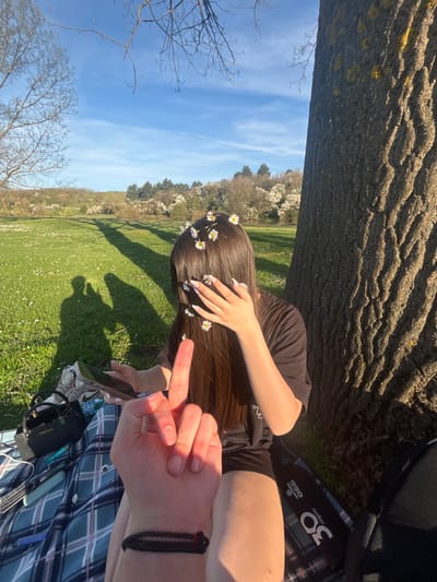 The image shows a partially obscured view of a woman lying in a grassy field in Beli Brod, Bulgaria. She is leaning against a tree trunk, with her head adorned with daisies. Her brown hair is spread out, and her right hand is raised near her face. She is wearing a dark-colored t-shirt.

In the foreground, a hand with a black bracelet is visible, interacting with the scene. A plaid blanket is spread on the grass, and various items, including a water bottle and a bag, are scattered around.

The background features a bright blue sky and a landscape with trees and vegetation. The lighting suggests it is daytime, possibly midday, with long shadows cast across the grass.