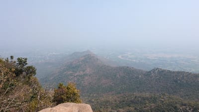 The image displays an elevated, panoramic landscape view over Kadaladi, India. In the immediate foreground, dense, mostly defoliated deciduous vegetation and a large, light-colored rock outcrop are visible. The midground features multiple undulating hills covered in sparse, dry, dark green and brown vegetation, with rocky outcroppings. A winding, brownish river is discernible in the distant right-center, flowing through a flat plain. Numerous clusters of small buildings, indicative of human settlements, are scattered across the plain in the far background. The sky presents a uniform, hazy light blue-gray, obscuring further distant details. No individuals, actions, or interactions are present in the scene.