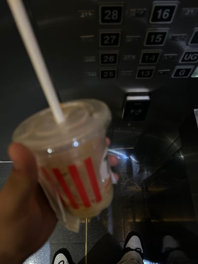 A first-person perspective shows a hand holding a plastic cup with a straw near an elevator panel with buttons labeled "16," "15," "14," "13," "28," "27," "26," and "25." The setting appears to be an elevator in a modern building. The floor is visible, with a person's shoes in the background. The general location is inferred to be in the city of Putrajaya, Malaysia.