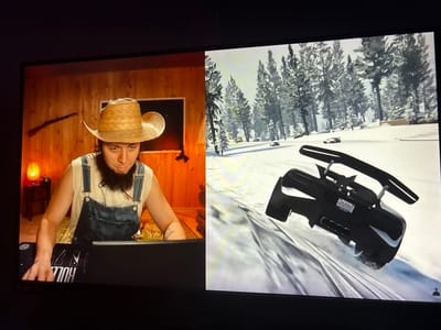 The image displays a split screen. The left side shows a person wearing a straw cowboy hat and denim overalls, sitting in front of a laptop. The background on the left is a rustic wooden wall with a lamp and a rifle mounted on it. The right side of the screen shows a video game scene with snow-covered pine trees and a black sports car on a snowy road. The car has a license plate that reads "MAJESTIC." The scene appears to be set in a wintery, mountainous environment. There is no information to suggest the location is the city of Kuvshinovo, Russia.