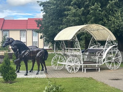 In the city of Капыльскі сельскі Савет, Belarus, a decorative scene features two dark bronze horse sculptures standing on a tiled pathway in front of a building with a red roof. The horses are positioned side-by-side, with the left horse slightly forward. To the right of the horses is a white wrought-iron carriage with a cream-colored canopy. The carriage has ornate scrollwork on its sides and large, decorative wheels. It appears to be an ornamental piece rather than a functional vehicle. The environment includes lush green grass in the foreground and background, mature trees, and a cloudy sky. The building in the background is a two-story structure with light-colored walls and multiple windows.