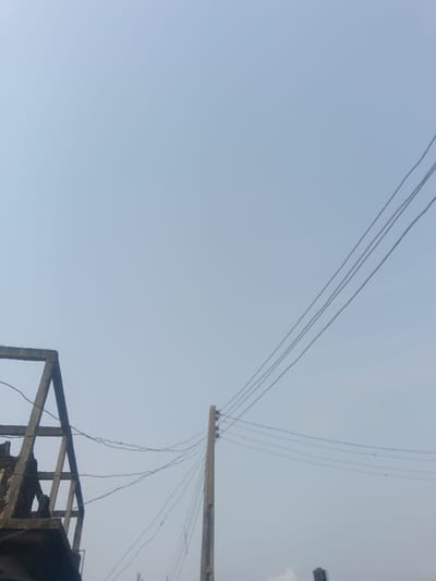 An upward-angled view in Okere-Urhobo, Nigeria, depicts an overhead scene under a light blue, hazy sky. In the lower left quadrant, the concrete skeletal frame of a structure, possibly an incomplete or derelict building, is partially visible, featuring exposed beams and columns. To the right of this structure, a utility pole, likely made of concrete, stands vertically, supporting multiple strands of electrical wiring via insulators near its apex. These wires extend diagonally upwards and across the frame. No individuals or specific activities are observable within the scene.