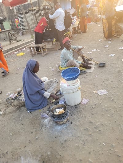 Several individuals are positioned outdoors on a dirt ground. One person in a light blue hijab kneels near a small cooking fire. Beside them is a bucket with a blue basket resting on top. An older man with a red hat sits crossed-legged and barefoot, holding a long object. More people, some wearing white and black clothing, are partially visible in the background. The setting appears to be a busy area, possibly a market or street.