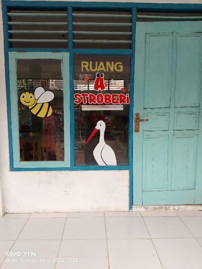 An exterior view depicts a building entrance in Jagapura Kulon, Indonesia, featuring a light blue-painted window adjacent to a light blue-painted wooden door. The window consists of two main glass panes with horizontal louvered sections above. On the left window pane, a cartoon-style yellow and black bee graphic is adhered. The right window pane displays yellow text reading "RUANG" at the top, a stylized red letter "A" with small red heart graphics, red and white text reading "STROBERI" below it, and a white stork cutout with a red beak at the bottom. Blurred interior elements are visible through the window glass. The wooden door to the right has a paneled design and is equipped with a brass-colored handle set. The building's exterior wall beneath the window and to its left is painted white, exhibiting minor discoloration at its base. The ground in the immediate foreground consists of light-colored square ceramic tiles. No people are visible.
