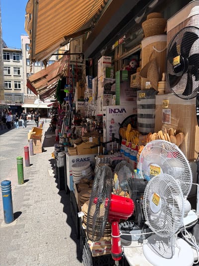 This image captures a bustling street market scene in Kadıköy, Turkey. The foreground is dominated by a display of various household appliances, primarily fans, stacked on shelves and the ground. A large black fan with a wooden-effect casing is prominently featured at the top left, while several white oscillating fans, some with price tags indicating "1.975" and "1.750", are arranged in the center. A bright red electric fan stands out amongst the collection. Other objects include woven baskets, what appear to be kitchen utensils like wooden spoons, and boxes of various products, some with brand names like "INOVA" and "RAKS".
In the background, awnings of shops cast striped shadows on the cobblestone street. Several people are visible in the middle ground, appearing to browse the market or engage in conversations, some wearing masks. The overall atmosphere suggests a daytime market, with bright sunlight creating distinct shadows. The presence of residential buildings with windows in the background hints at a mixed-use urban environment. The assortment of goods and the casual arrangement suggest a lively and somewhat unorganized retail setting, typical of many street markets. The visible text includes prices, brand names, and product descriptions in Turkish.
